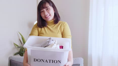 Young-Asian-woman-looking-at-camera-holding-box-with-clothes-for-donation