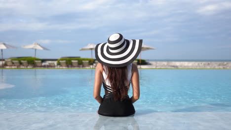 Toma-Estática-De-La-Espalda-De-Una-Mujer-Elegante-Con-Sombrero-Blanco-Y-Negro-Y-Traje-De-Baño-Sentado-En-La-Piscina-De-Un-Hotel-De-Lujo,-Espacio-Para-Copiar