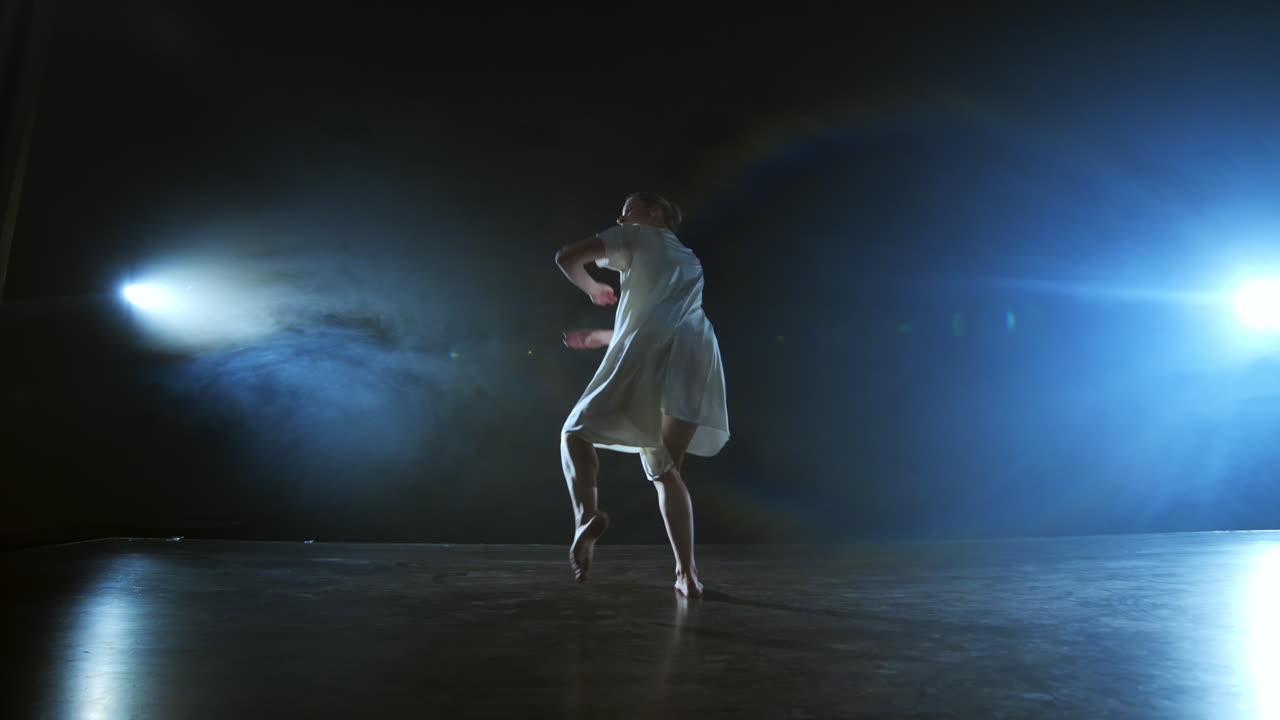 a young girl in a white dress dances a modern ballet makes rotations and jumps in slow motion on the stage with smoke against the spotlights.