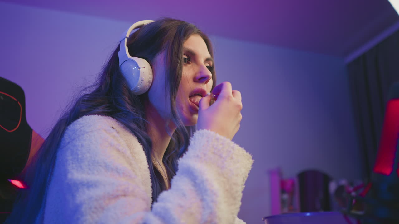 Side view of woman with headphones in cozy purple lit studio reaching into bowl of popcorn while working on computer surrounded by colorful lights and microphone in frame for streaming vibe