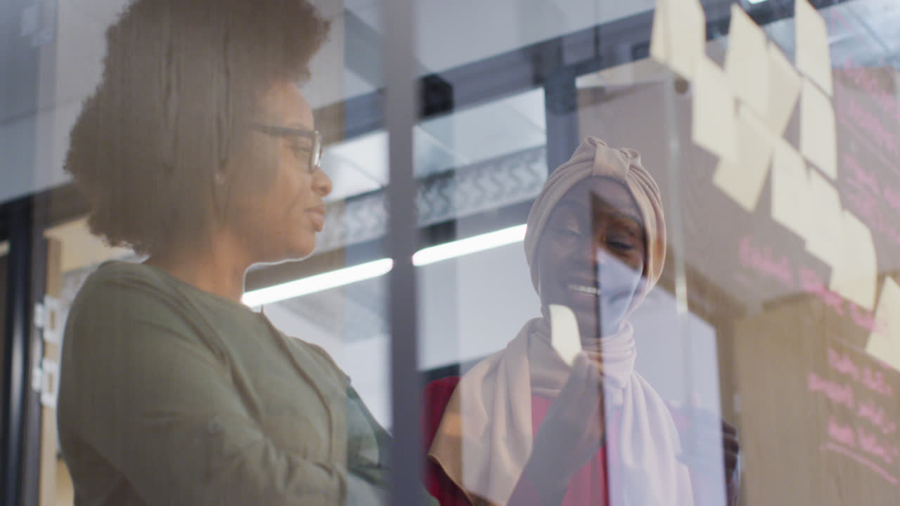 두 명의 미소 짓는 아프리카계 미국인 사업가 여성이 사무실의 유리 벽에 메모 노트를 사용하여 브레인 스토밍을합니다.