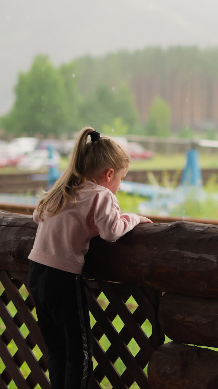una niña juguetona con una sudadera rosa se sube a la valla de troncos para ver operar un pequeño helicóptero en el prado en un hotel ecológico en un día nublado en cámara lenta