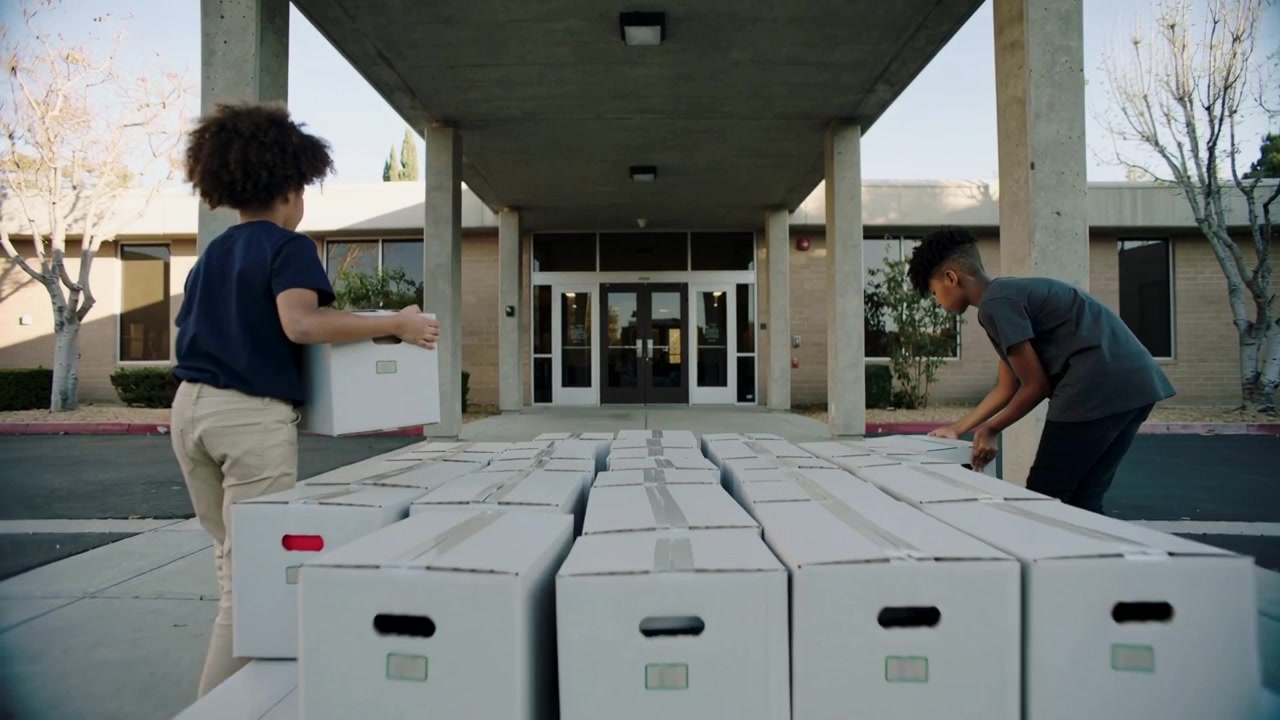 Starting boxes opposite, teens in casual tees stacking white corrugated boxes forming entrance wall