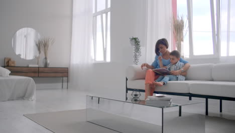 a young mother with a child reading a book sitting in a bright white interior of the house in the living room on the couch