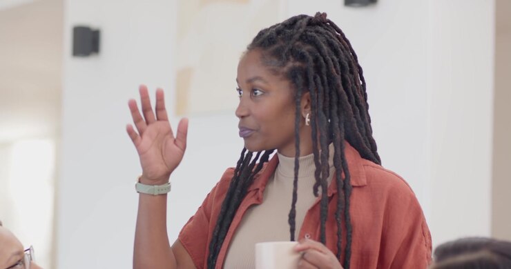 Diverse team watching standing woman holding mug, explaining after Asian peer gesturing in office