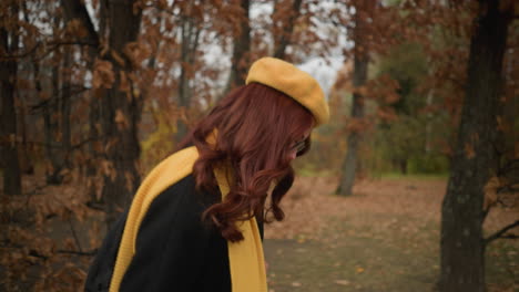 woman with yellow muffler and beret walks through forest, looks up and down as wind blows her red hair back, surrounded by autumn leaves