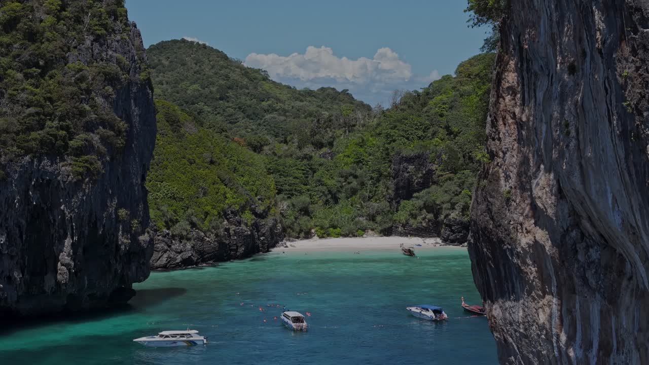 Drone pans left revealing a hidden white sand beach and crystal-clear turquoise bay, surrounded by cliffs and lush greenery in Phi Phi Islands, Thailand.