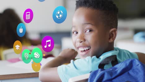 Animation-of-school-icons-over-happy-african-american-schoolboy-sitting-at-desk-in-class