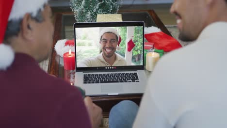 Padre-E-Hijo-Birraciales-Sonrientes-Usando-Una-Computadora-Portátil-Para-Una-Videollamada-Navideña-Con-Un-Hombre-En-La-Pantalla