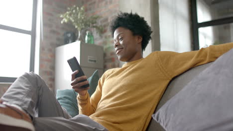 happy african american man sitting on sofa using smartphone at home, slow motion