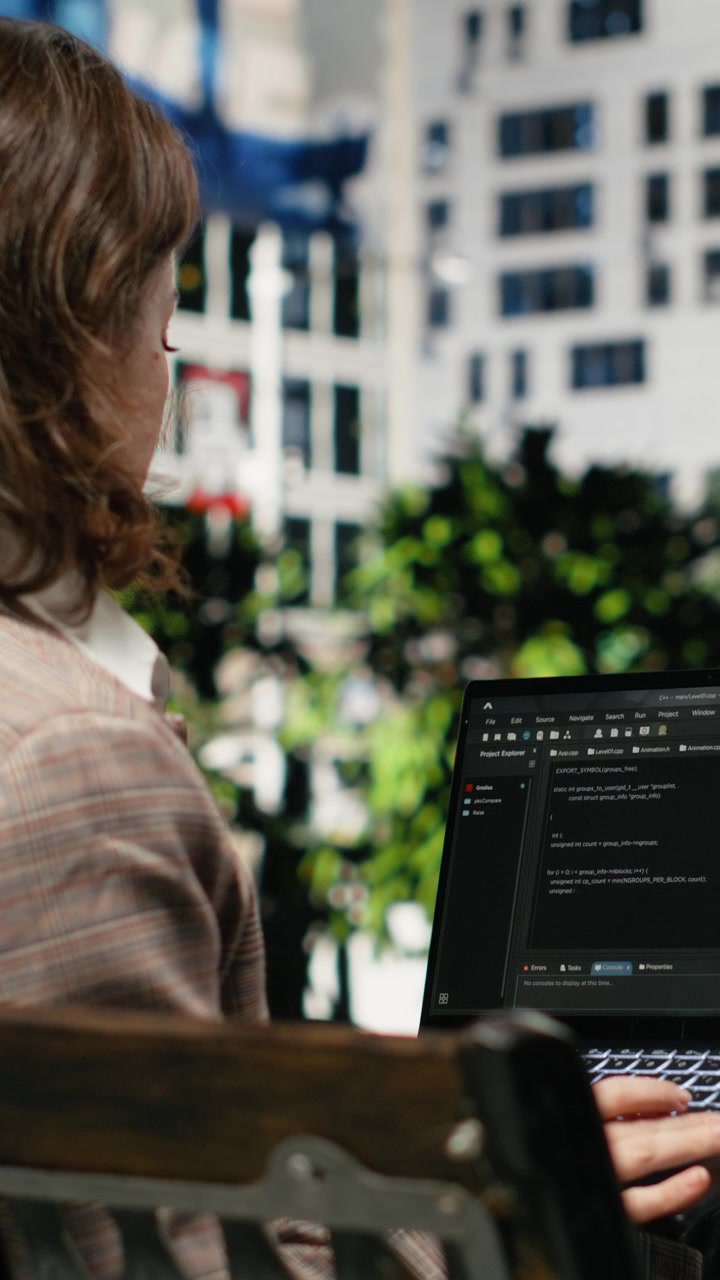 Vídeo vertical: Vista traseira de programadora em banco de parque escrevendo código no laptop.