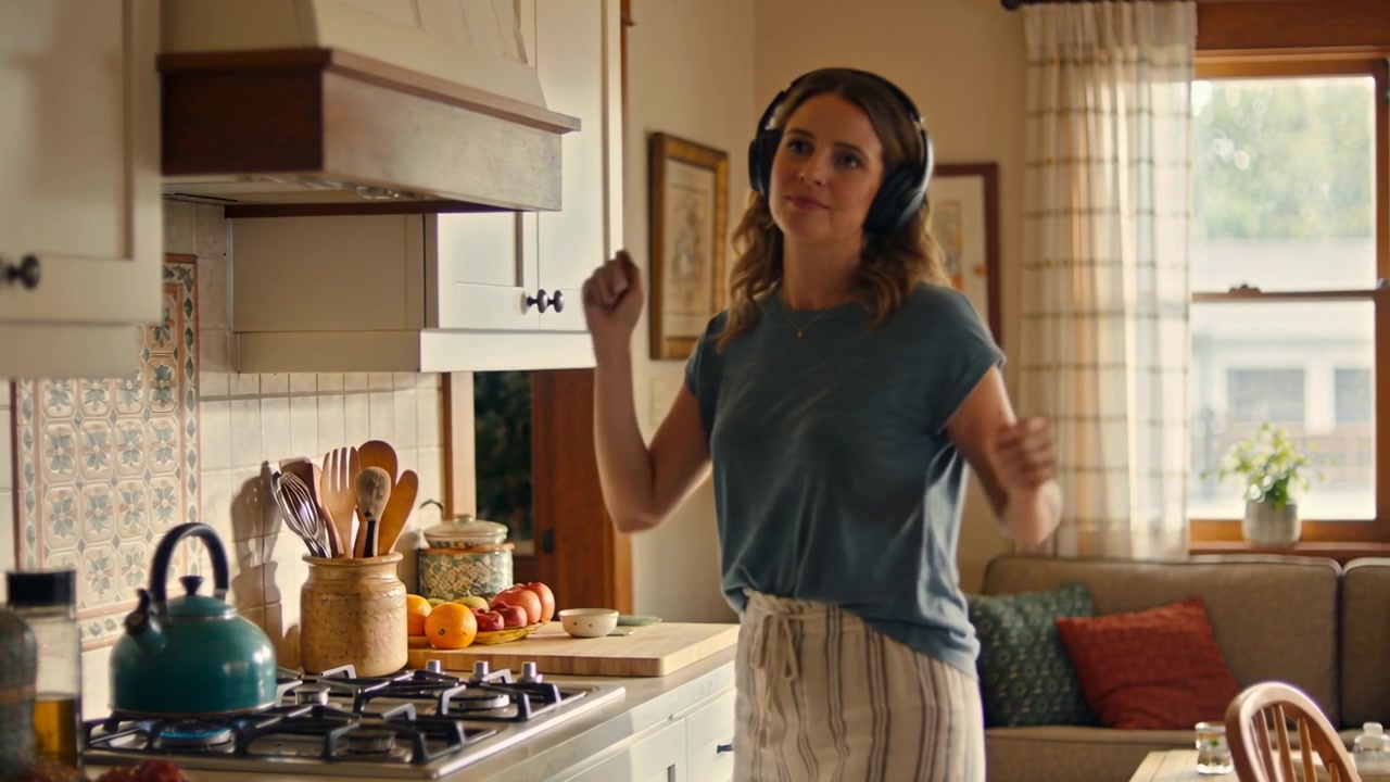 Donna che balla con un top blu, ascoltando musica, si gira verso la telecamera al bancone della cucina con un bollitore color verde acqua.