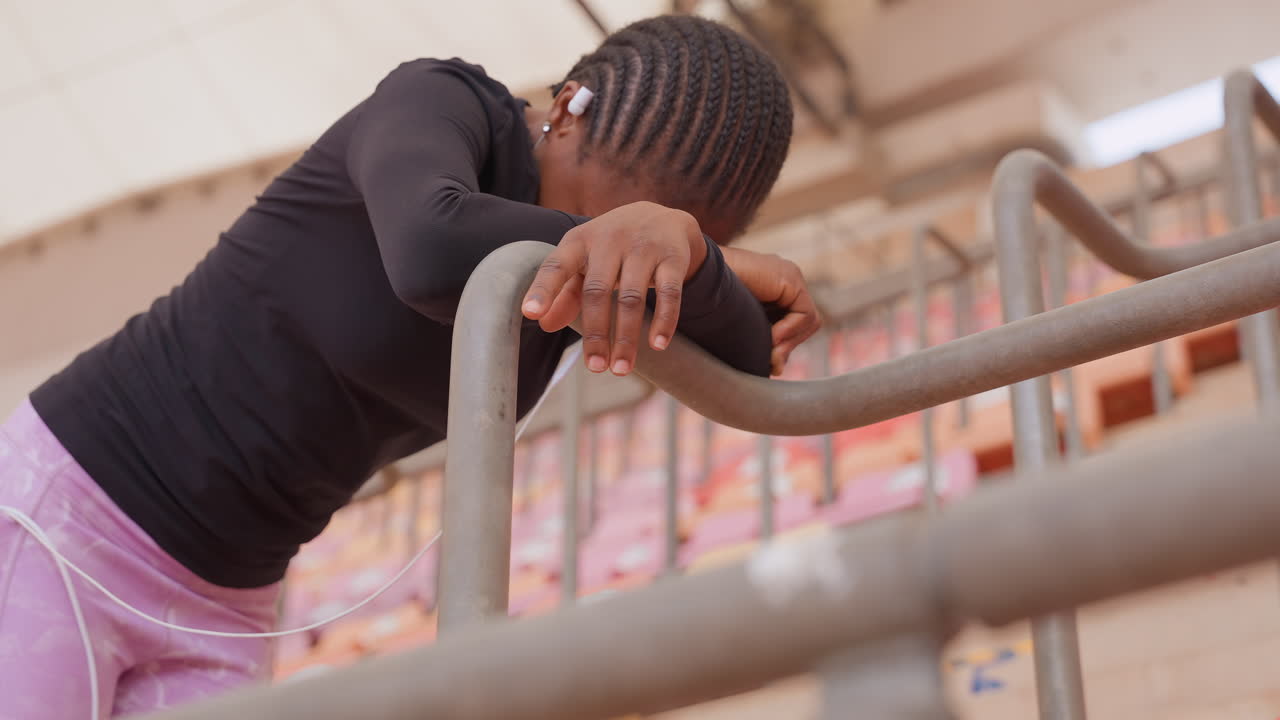 Exhausted woman leaning over metal railing, hands gripping rail and pink leggings visible, head bowed into forearm with earbuds in, intimate view of vulnerability after intense workout in empty arena