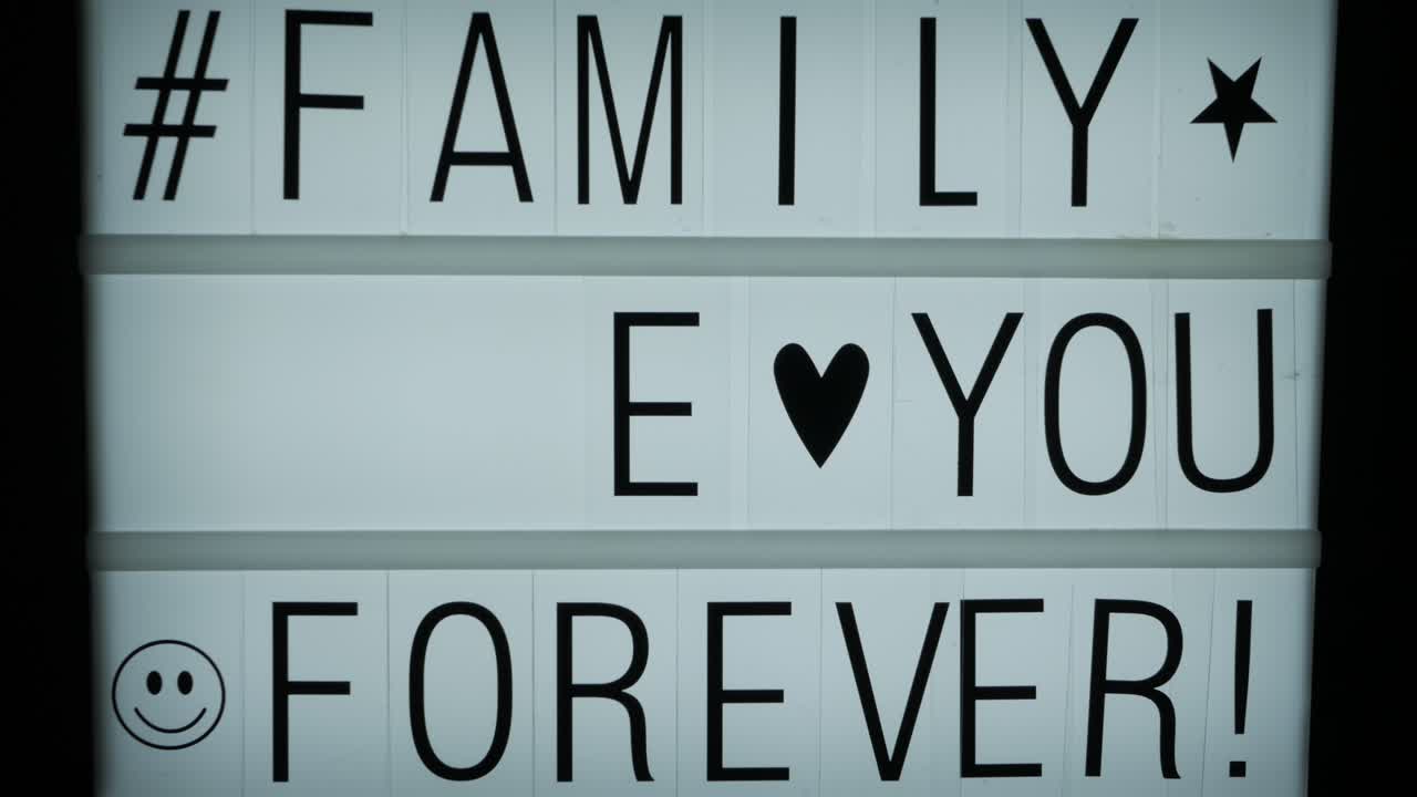 una mano completando un mensaje de texto en una cartelera retroiluminada: la familia te ama para siempre, un hashtag y algunos símbolos