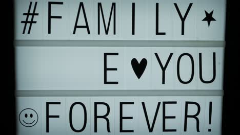 a hand completing a text message on a backlit billboard: family love you forever, a hashtag and some symbols