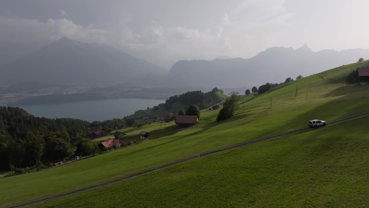 A white car travels along a narrow mountain road surrounded by green meadows, traditional houses, and a calm lake under soft daylight. Peaceful and adventurous alpine scenery