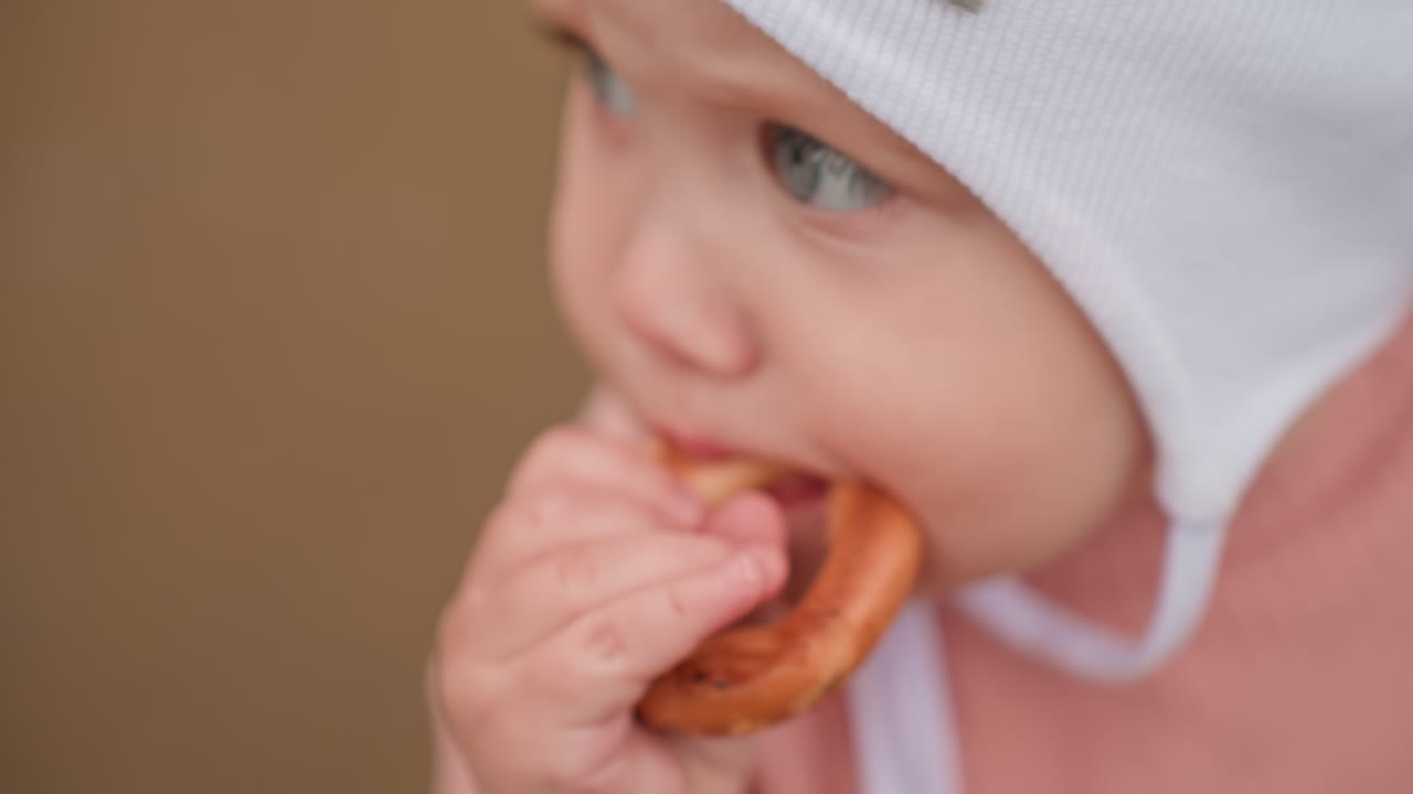 Close Up Baby Biting Pretzel, Small Hand Gripping Pastry And Determined Gaze, Wearing White Cap And Pink Sweater, Crunchy Snack Exploration During Stroller Outing, Candid Feeding Moment With Crumbs