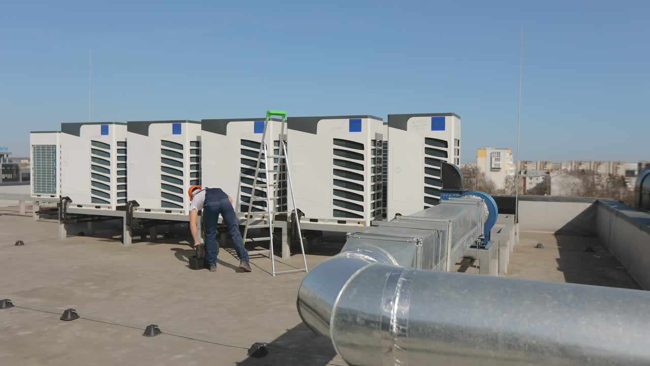 A young engineer is opening a toolbox and taking a pipe wrench. He's on the roof of a business center. A ladder is standing beside him. He is wearing work clothes and a hard hat. Nice sunny day. 4K
