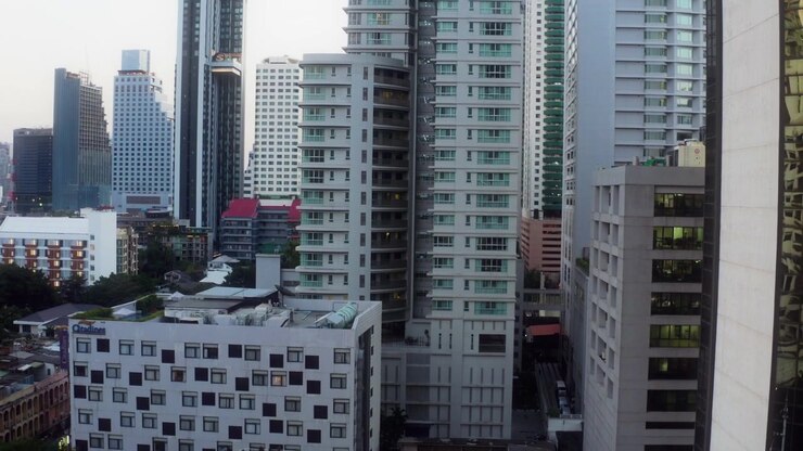 Drone desce verticalmente entre arranha-céus densos em Bangkok, revelando arquitetura urbana moderna e telhados abaixo.