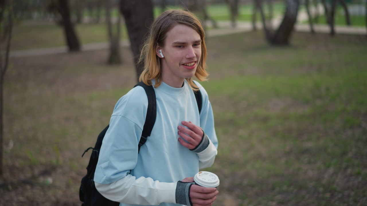 주변을 둘러보며 커피를 들고 있는 젊은이, 보이는 백팩 끈, 생각에 잠긴 시선, 밝은 스웨트셔츠, 천천히 마시는 모습, 부드러운 오후 햇살, 공원 통로, 나무, 편안한 걸음, 결정의 순간