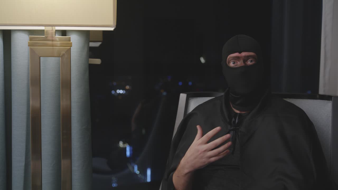 falso documental de superhéroes desempleados - hombre con máscara negra completa sentado y explicándose frente a la cámara