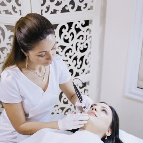 an elegant beautician in a lab coat talking to a client during a permanent lip makeup procedure on the background of white decorative wall at a beauty salon. close-up.