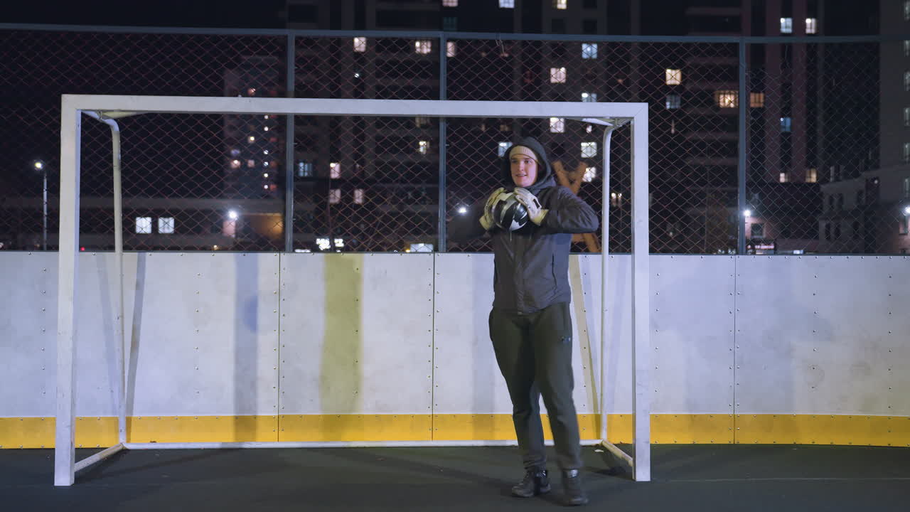Goalkeeper in athletic gear makes a precise catch to the left, stopping a powerful soccer ball shot on goal during an intense nighttime practice session in an urban outdoor field