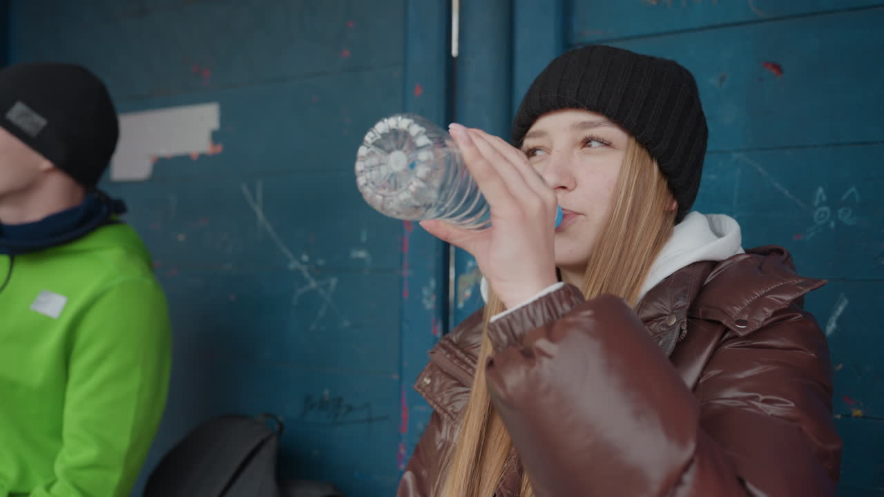 운동 후 야외 쉼터에서 생수를 마시는 백인 여성과 남성, 남성은 녹색 재킷을 입고 여성은 갈색 패딩을 입고 있으며, 옆에는 백팩과 장비가 있고, 안도의 숨을 쉬며 회복하는 도시의 모습