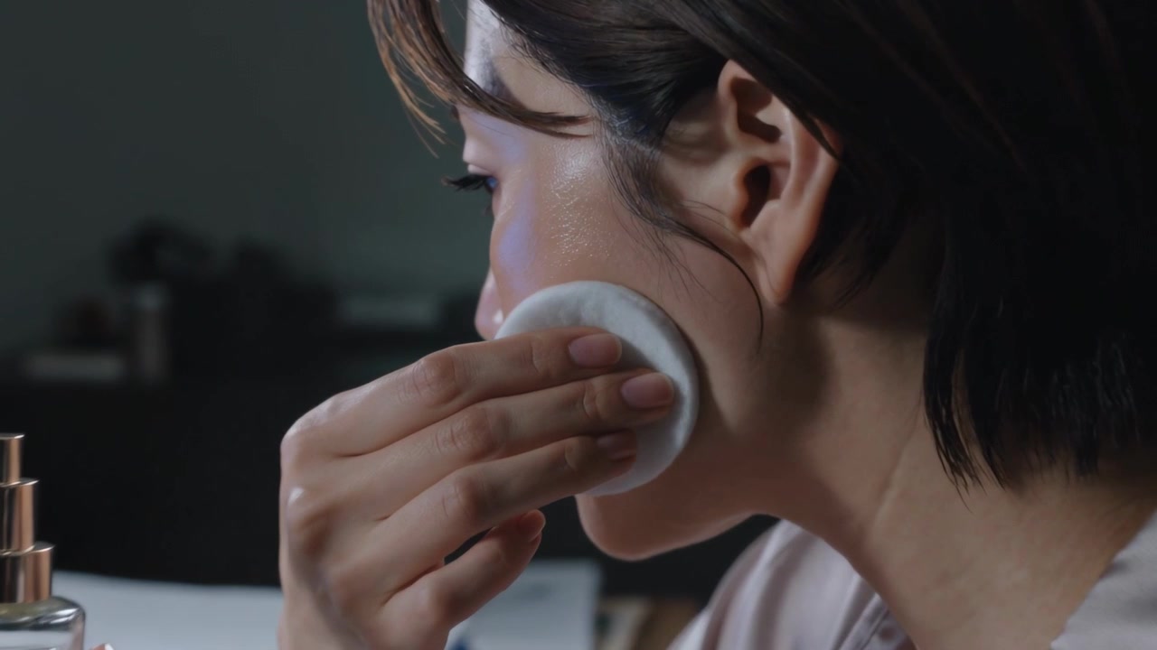 Pressing pump, Asian woman in satin blouse cleansing face at desk, using cotton pad and lamp
