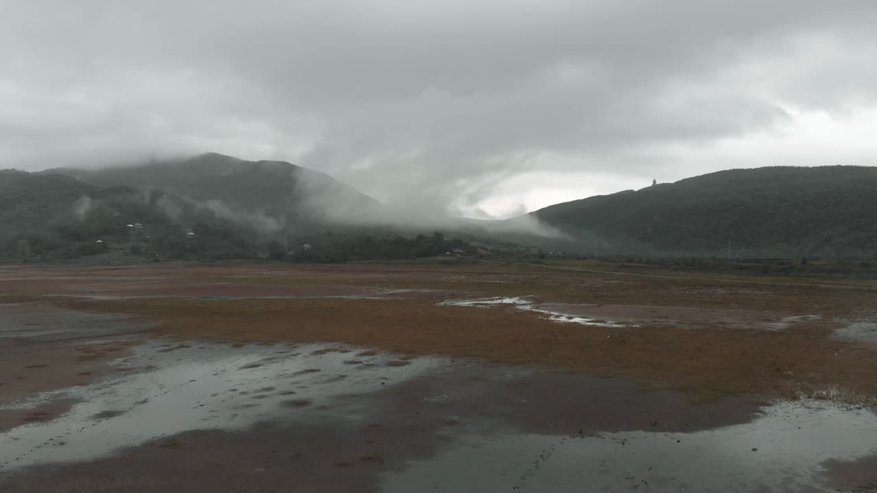 un dron volando bajo sobre una playa húmeda que se eleva hacia las montañas cubiertas de nubes en la distancia