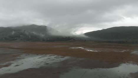 un dron volando bajo sobre una playa húmeda que se eleva hacia las montañas cubiertas de nubes en la distancia