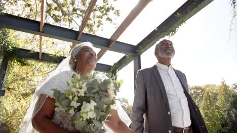 Happy-senior-biracial-couple-walking,-holding-hands-during-wedding-ceremony-in-garden,-slow-motion