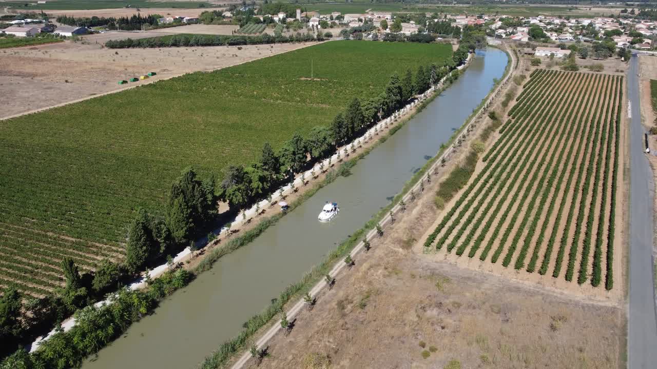 Barca che lascia un villaggio su un canale, pista pedonale e ciclabile sulla riva con rigogliosi vigneti verdi, fine estate nel Sud della Francia