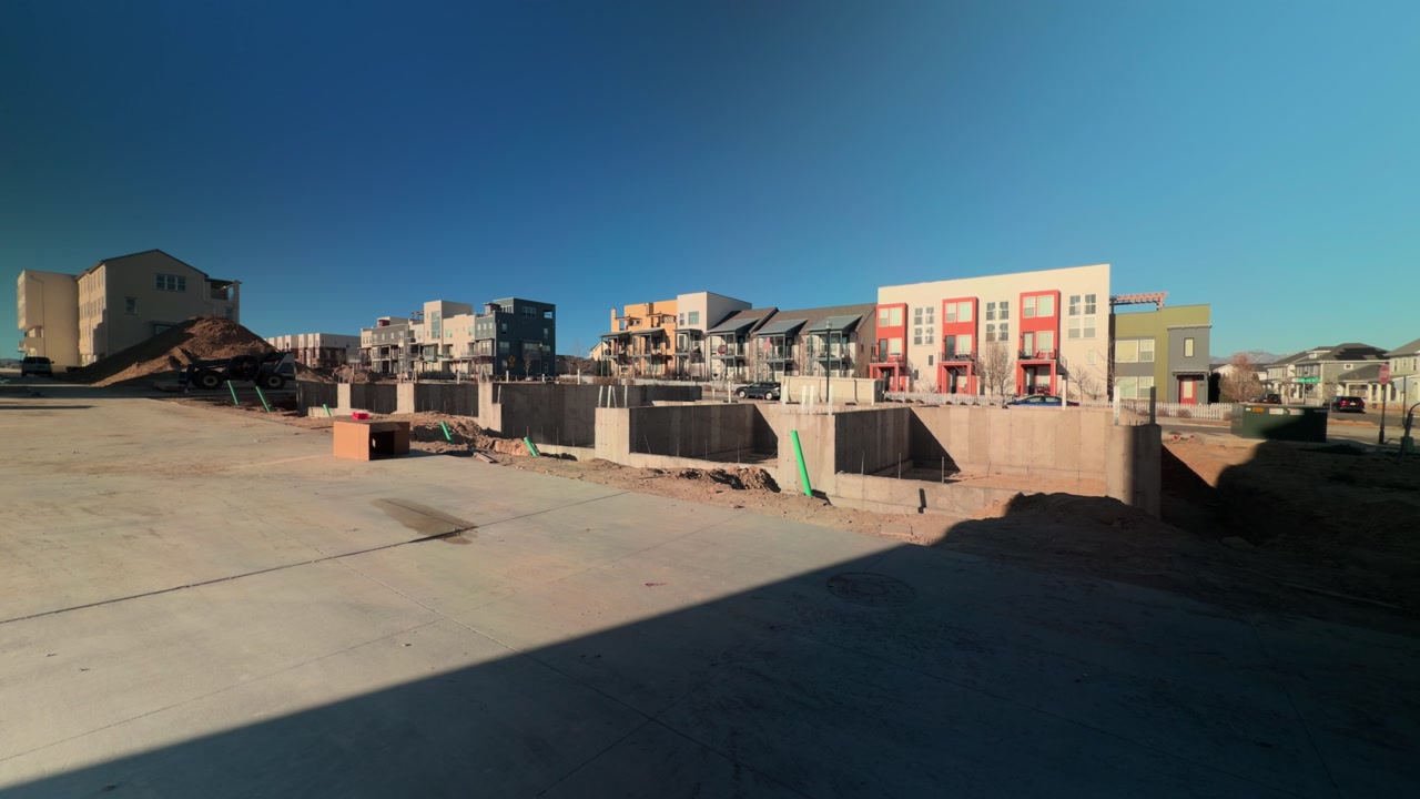 Wide angle slow pan across foundation walls, graded site, and finished housing beyond.