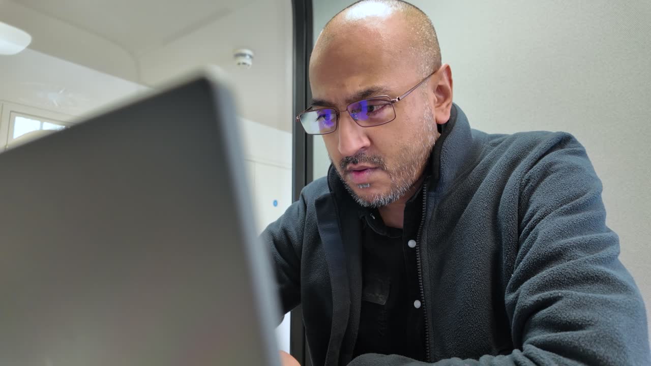 South Asian male office worker concentrating deeply while using a laptop in a sleek, modern office pod, enhancing productivity and focus