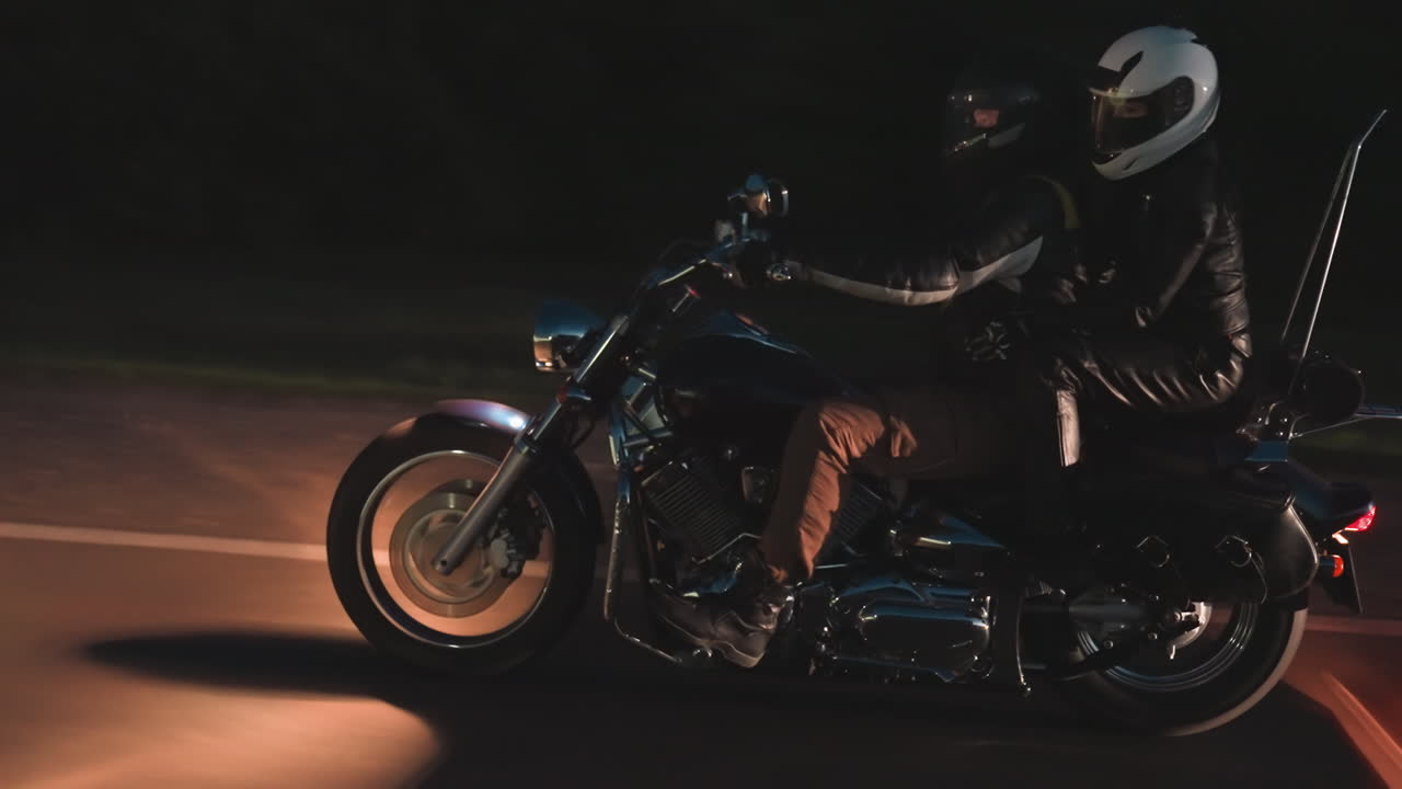 Motorcyclist with passenger rides at night on dark road, headlights illuminating path while car follows from behind, creating reflective glow on chrome bike parts and asphalt surface