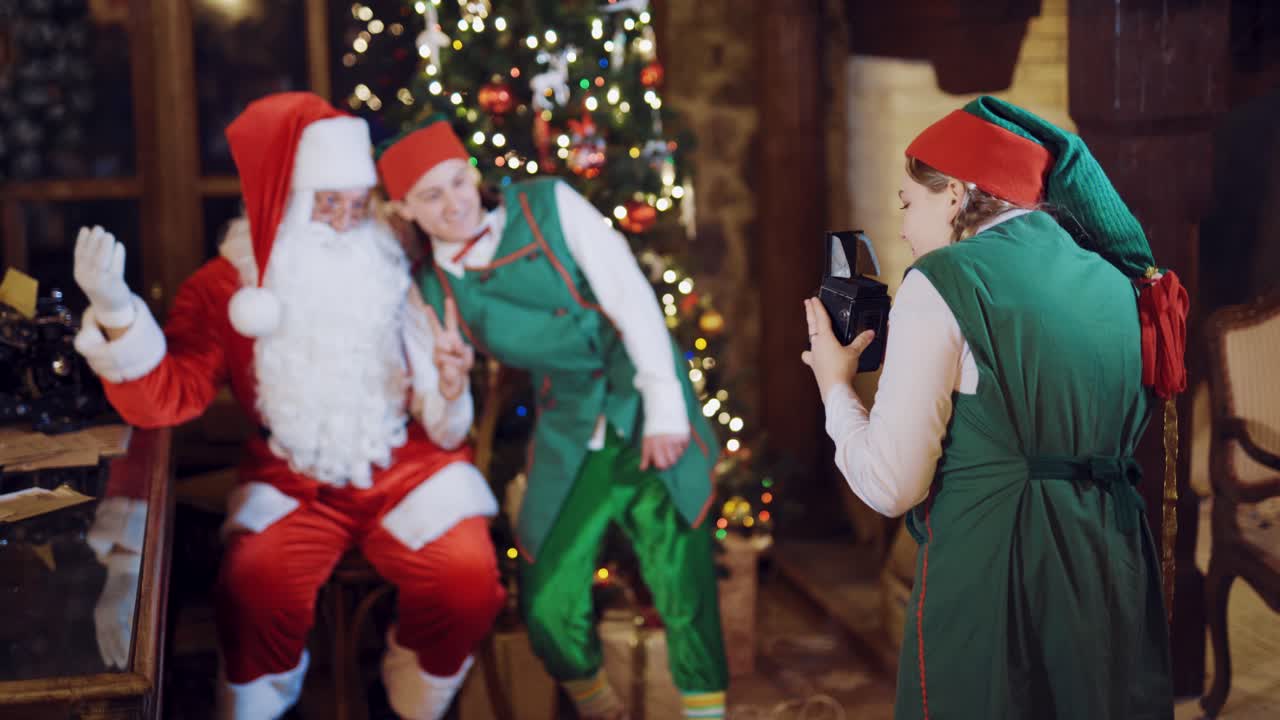 An assistant elf is photographing Santa Claus and an elf on the background of a Christmas tree and garlands near the window using a retro camera on New Year's Eve. Blurred background.