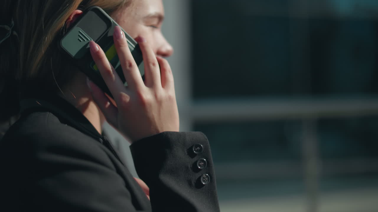 Close up side view of young girl in formal outfit laughing joyfully while on phone call with friend outside in sunlight, background featuring modern glass building with soft blur