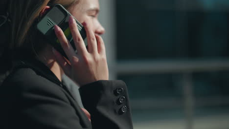 close up side view of young girl in formal outfit laughing joyfully while on phone call with friend outside in sunlight, background featuring modern glass building with soft blur