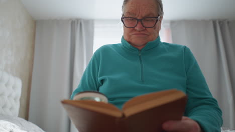 elderly woman sitting on bed, holding open book in hands with magnifying glass. she seems focused on reading, possibly struggling with small print or inspecting details in the text