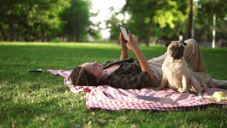 woman laying on plaid on lawn in a park and reading e-book while little pug sitting next to her