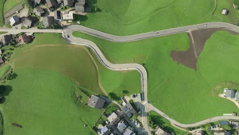 Overhead-aerial-tilt-down-view-of-cars-moving-on-a-winding-road-of-the-small-village-of-La-Val,-South-Tyrol,-Italy-with-it's-lush-green-steep-grass-covered-hills