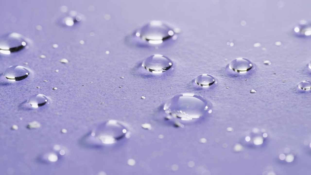Primo piano di goccioline d'acqua limpide di varie dimensioni che poggiano su carta lavanda testurizzata; le goccioline mantengono forme a cupola lisce con riflessi speculari nitidi e riflessi neutri, mentre minuscoli elementi metallici