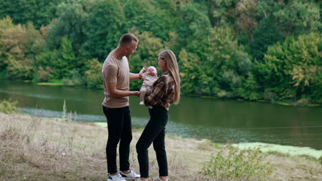 beautiful caucasian family of three standing in the nature at the river bank. parents of newborn look at their child and then kiss.