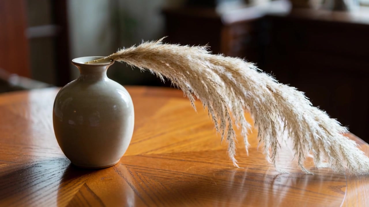 Swaying pampas plumes drifting from pale ceramic vase across round wooden table, draft from window