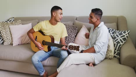 Smiling-mixed-race-gay-male-couple-sitting-on-sofa-one-man-playing-guitar