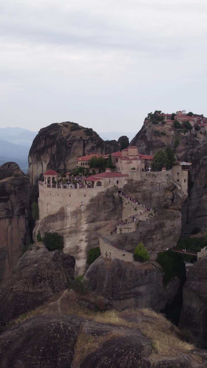 Ripresa aerea verticale ad arco che cattura il monastero di Varlaam in cima ai pilastri di roccia delle Meteore, rivelandone la drammatica elevazione e il paesaggio circostante