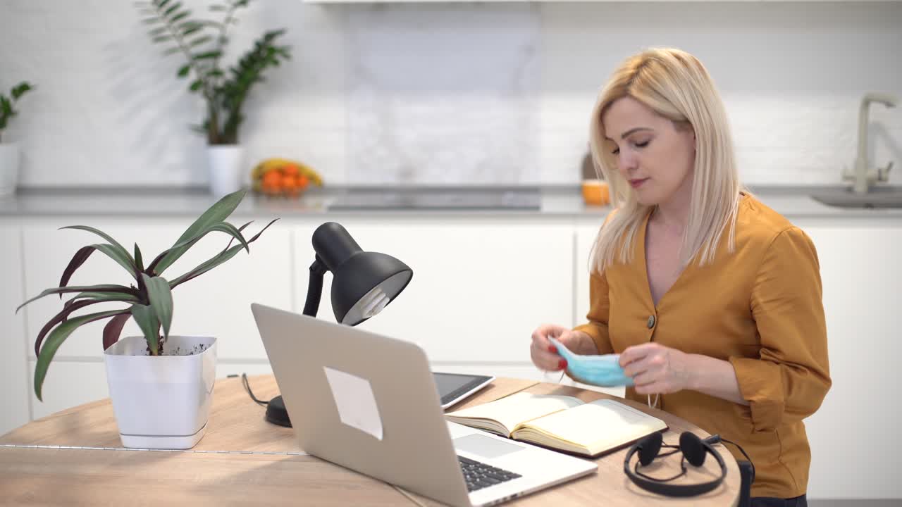 Kvinde med beskyttelsesmaske arbejder hjemmefra på bærbar computer i køkkenet.