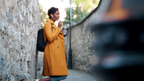 Young-adult-woman-wearing-a-yellow-pea-coat-talking-using-her-smartphone-earphones-and-drinking-a-takeaway-coffee,-leaning-on-a-stone-wall-in-a-historical-alleyway,-low-angle,-rack-focus