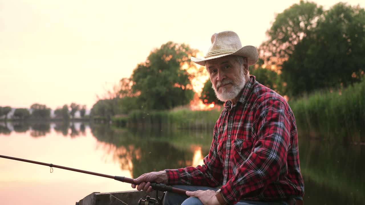 Portrait of a senior fisherman putting on hat and sitting on the lake pier while holding a fishing rod at sunset